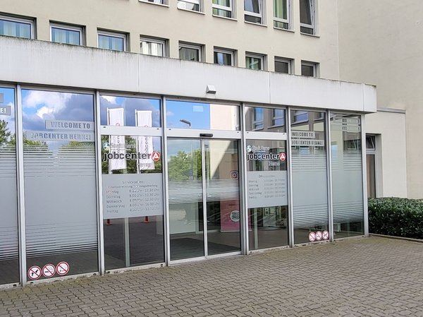 Eine Frontalaufnahme des Haupteinganges des jobcenter Herne in der Koniner Straße in Herne. Man sieht die gläsernen Windfang.